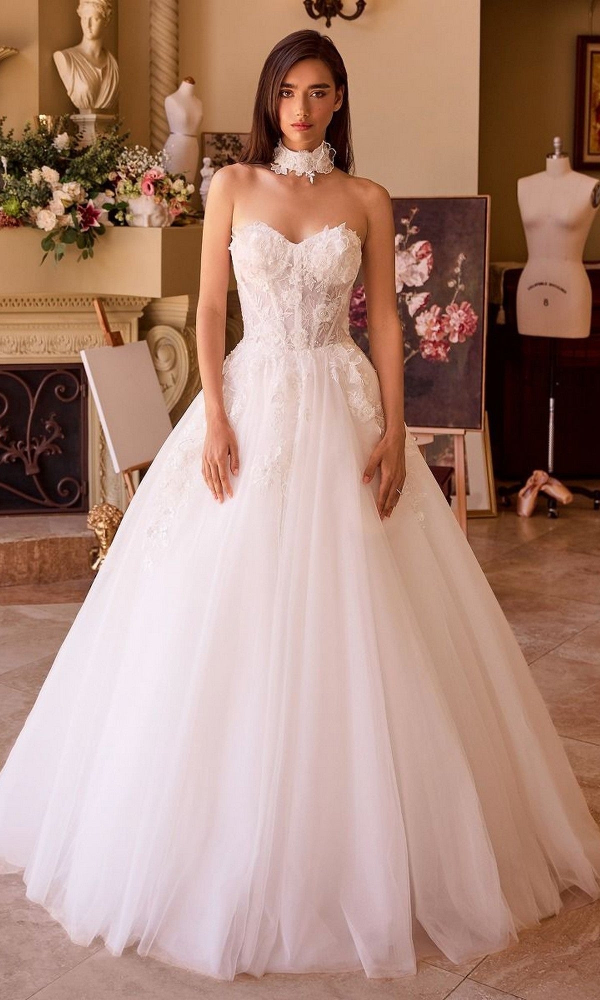 A woman stands indoors wearing the Andrea and Leo WL041 Long Wedding Dress, which features a strapless floral lace bodice and an A-line tulle skirt. Art supplies and a floral arrangement are visible in the background.